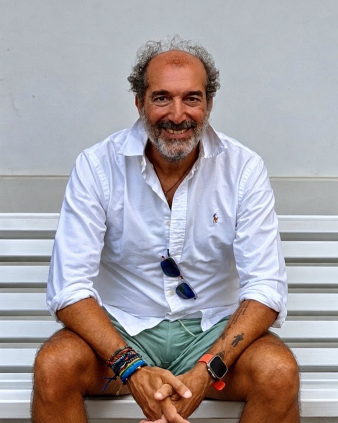 Pablo Navarro, con camisa blanca, sonríe sentado en un banco delante de una pared clara